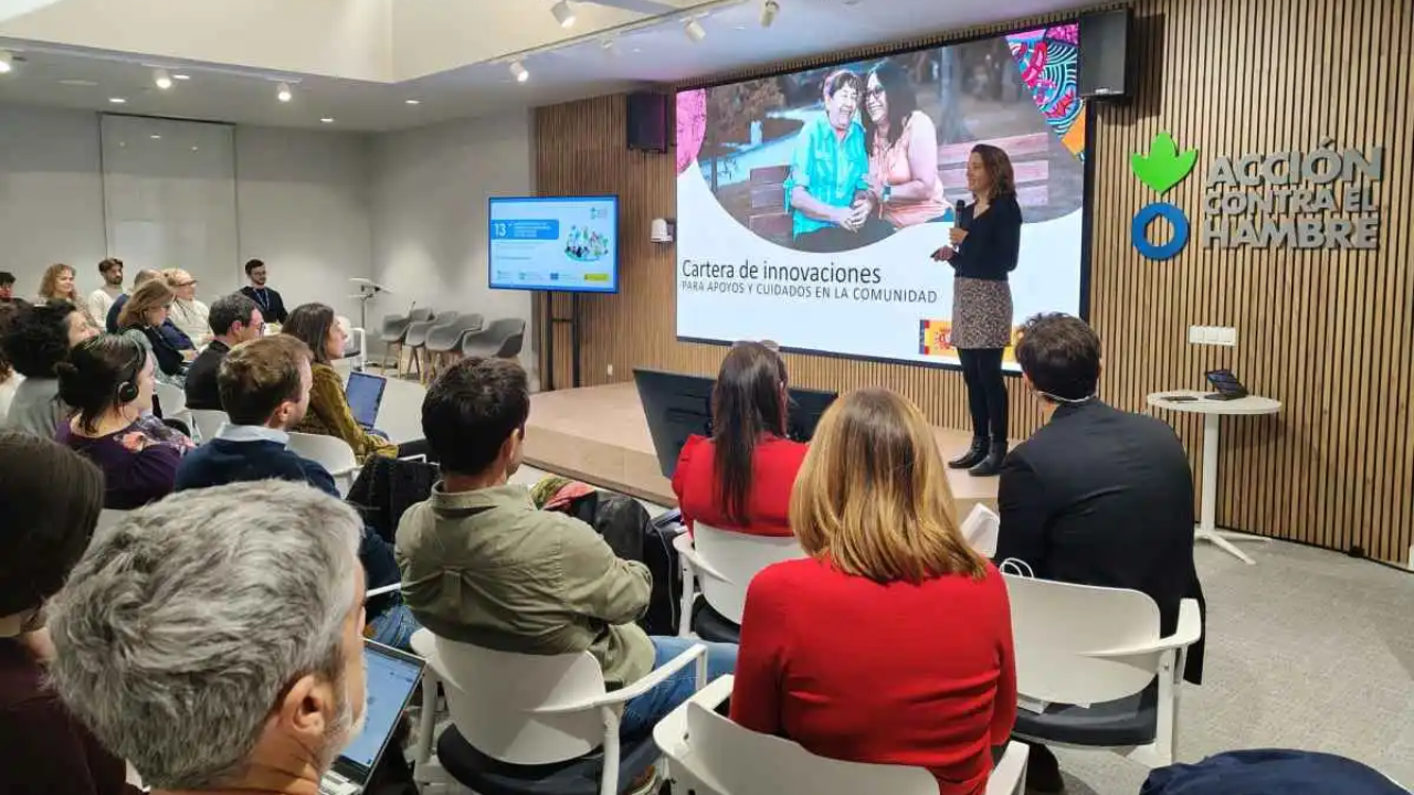 Un grupo de personas sentadas escucha a una mujer hablando en un escenario con una pantalla detrás