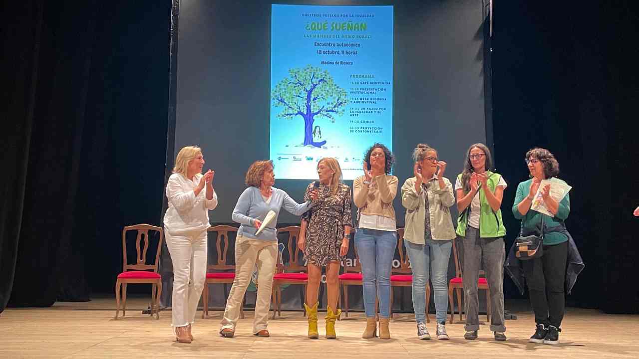 Siete mujeres aplauden sobre un escenario con una foto de un árbol detrás
