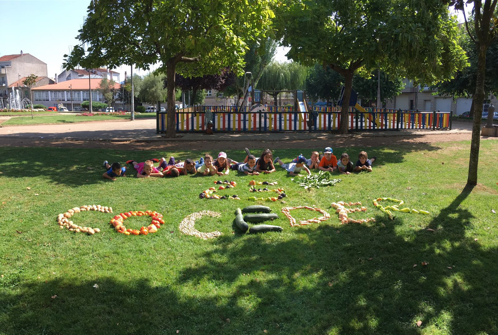 Grupo de niños tumbados en un cesped con la palabra coceder realizada con frutas y verduras