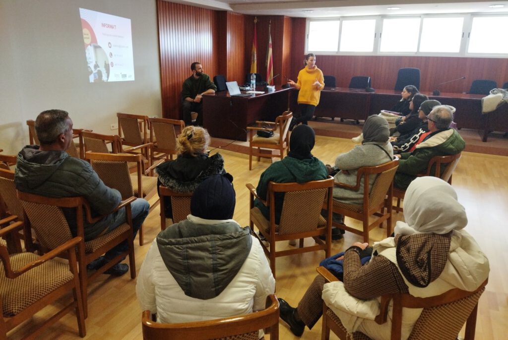 grupo de personas en un salón escuchando la información ofrecida por dos personas, una en una mesa y otra en pie gesticulando