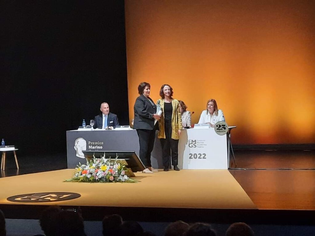 Atril con personas presentando los premios y en pie dos mujeres esperando la entrega del mismo